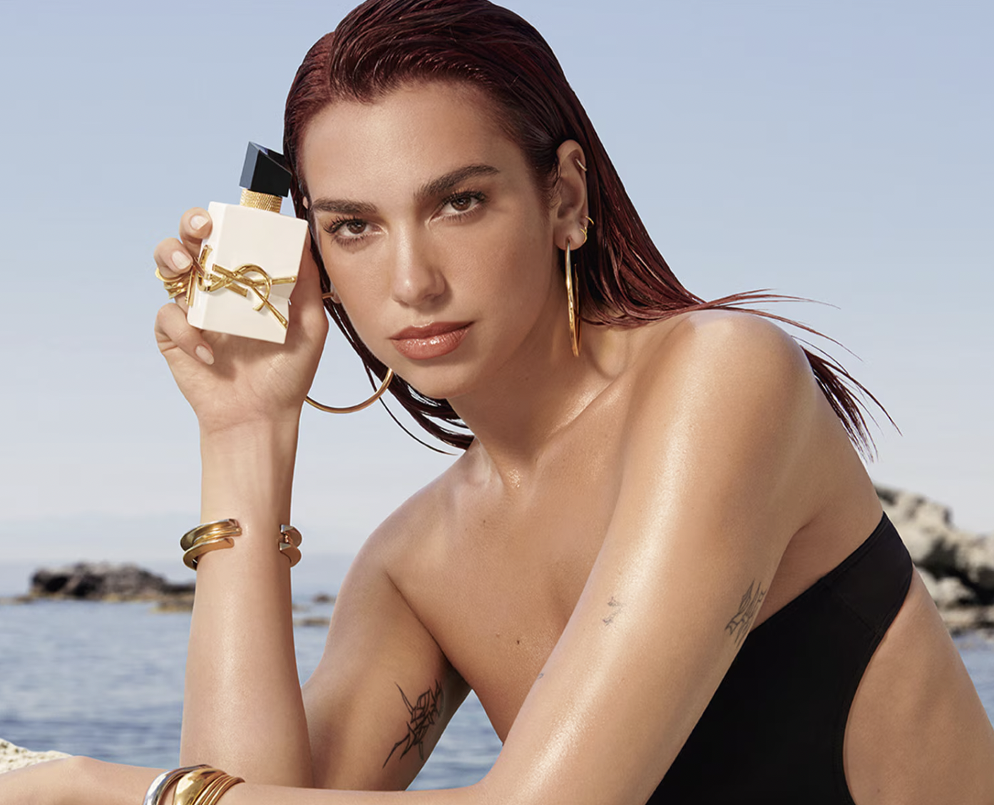 Dua Lipa posando junto a rocas frente al mar con el frasco de perfume, vistiendo un traje de baño negro.