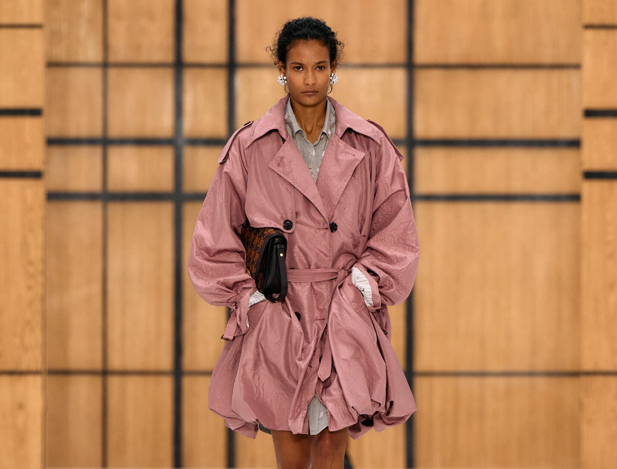 Modelo con un abrigo ligero rosa texturizado estilo trench, camisa y vestido interior gris, con un bolso de mano oscuro y tacones beige