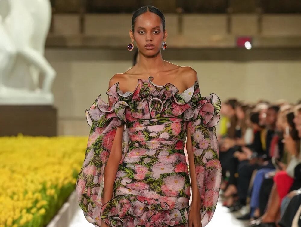 Modelo desfilando con un vestido corto de hombros descubiertos, mangas abullonadas y volantes, con un estampado floral en tonos rosados y oscuros.