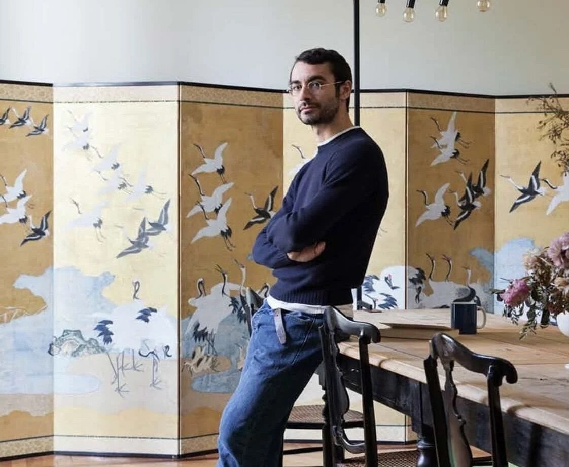 Dario Vitale de pie en un estudio con un biombo decorado con grullas doradas al fondo, vistiendo un suéter azul marino, jeans y tenis oscuros.