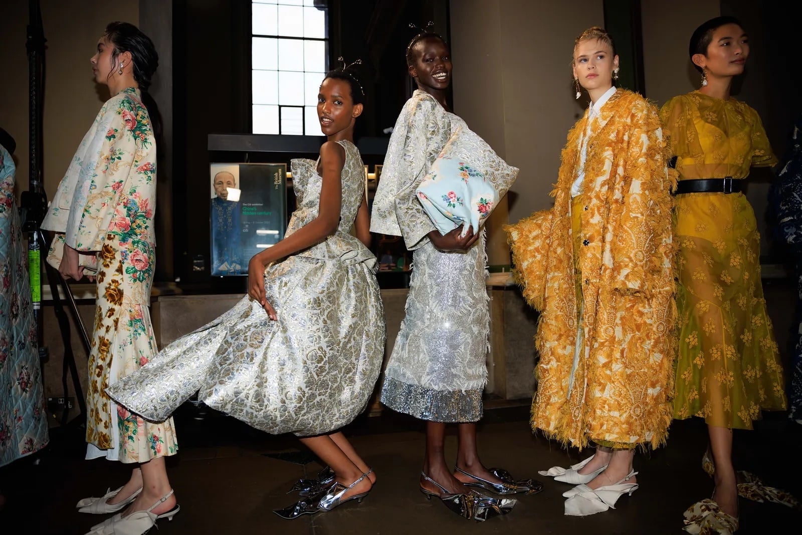 Grupo de modelos posando en backstage con abrigos largos estampados, vestidos fluidos de brocado y conjuntos texturizados en tonos dorados y blancos.