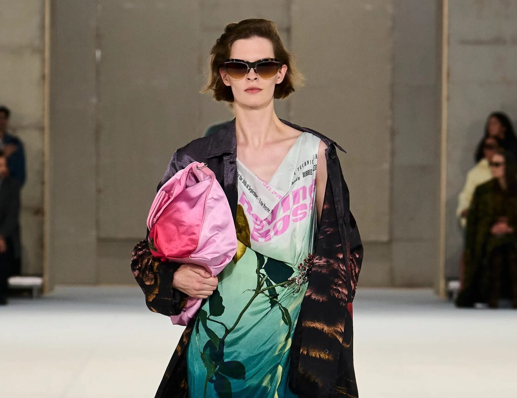 Modelo desfilando con un vestido midi estampado en tonos verdes y azules, cubierto por una gabardina larga oscura, gafas de sol y cargando un bolso rosa brillante.