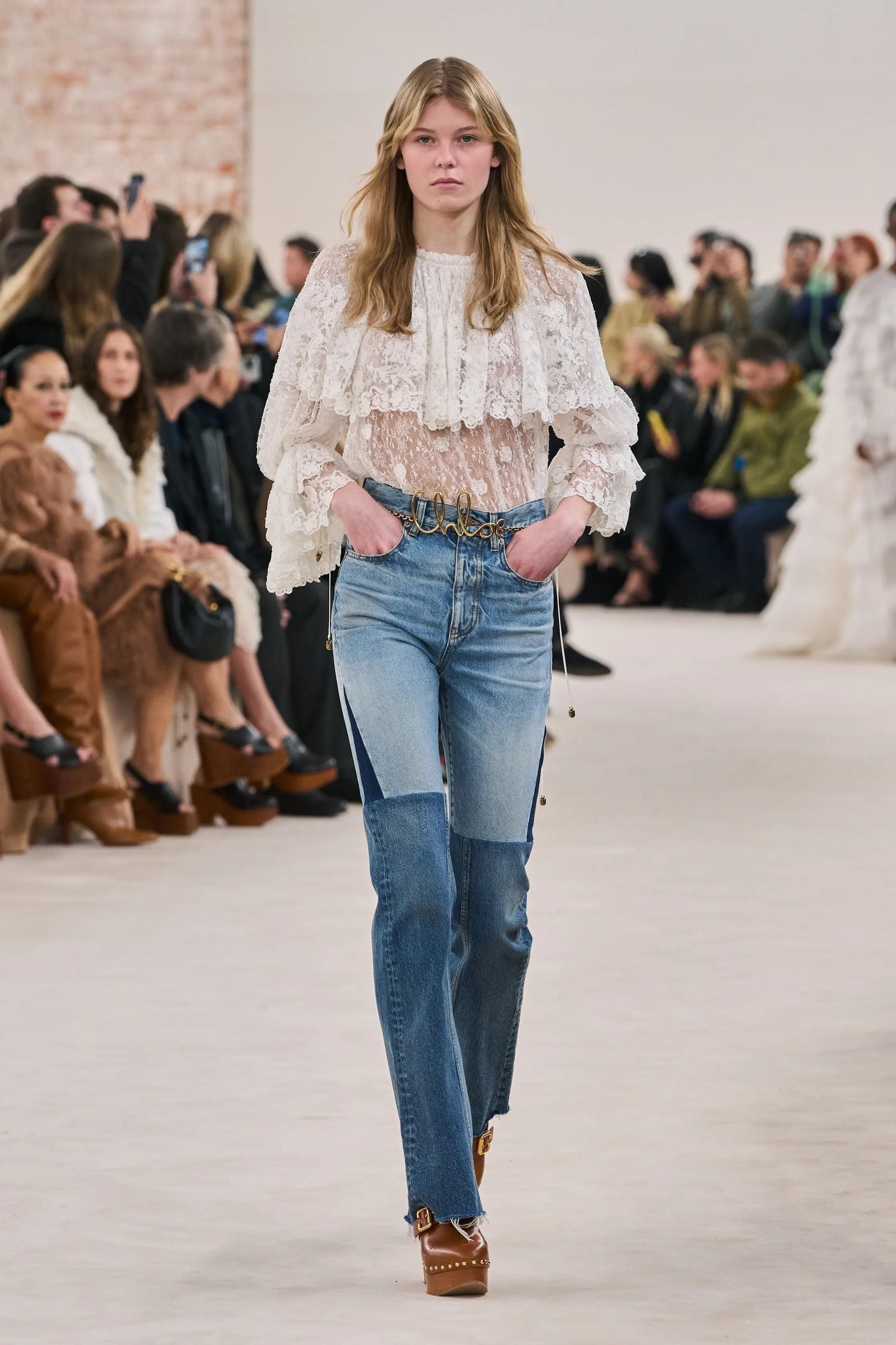 Modelo en desfile de moda luciendo jeans bootcut con paneles de distintos tonos de mezclilla, blusa romántica de encaje blanco con volantes y cinturón con hebilla metálica.