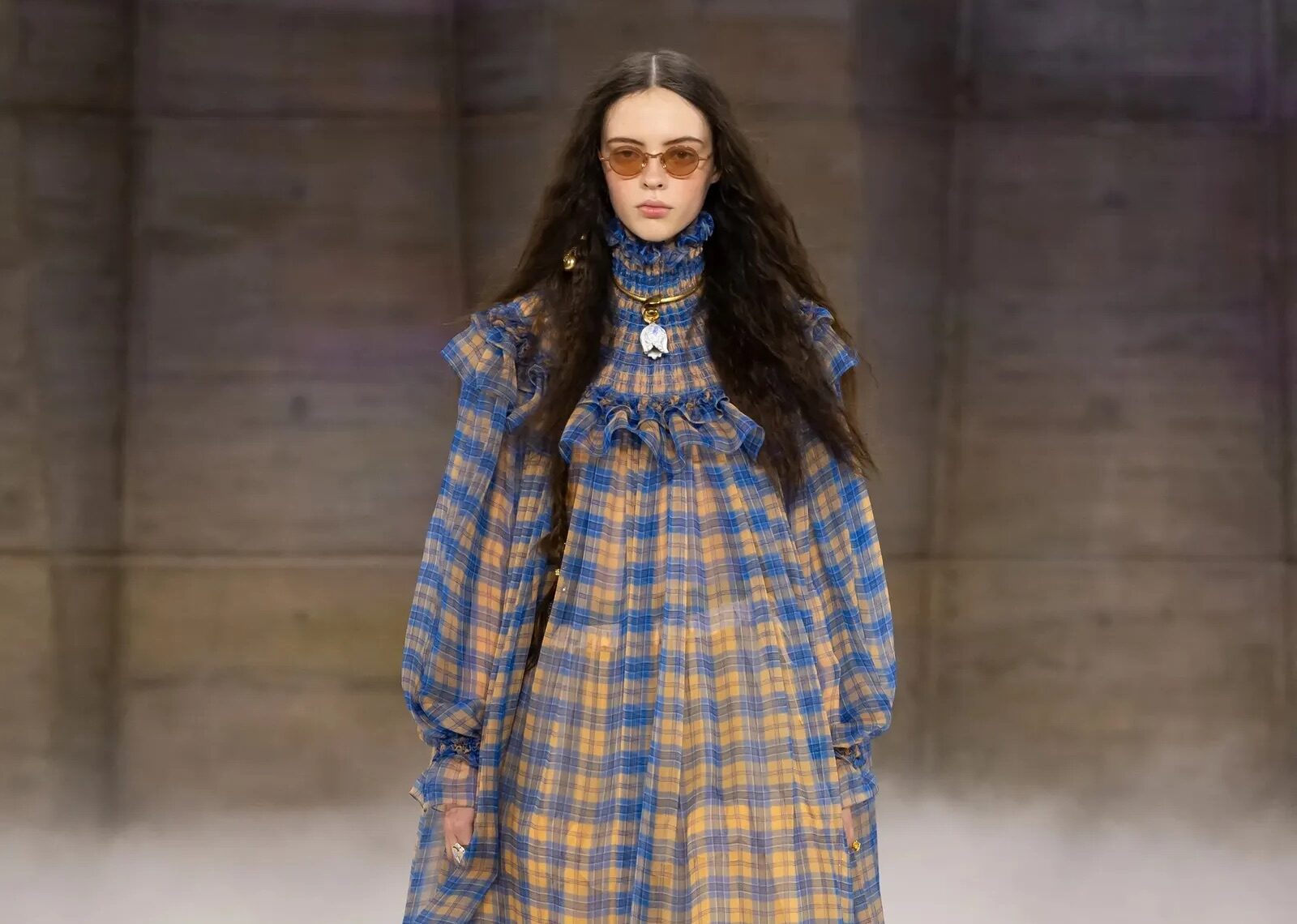Modelo en pasarela luciendo un vestido largo de cuadros en tonos azules y amarillos con volantes en el cuello y el dobladillo, combinado con botas altas.