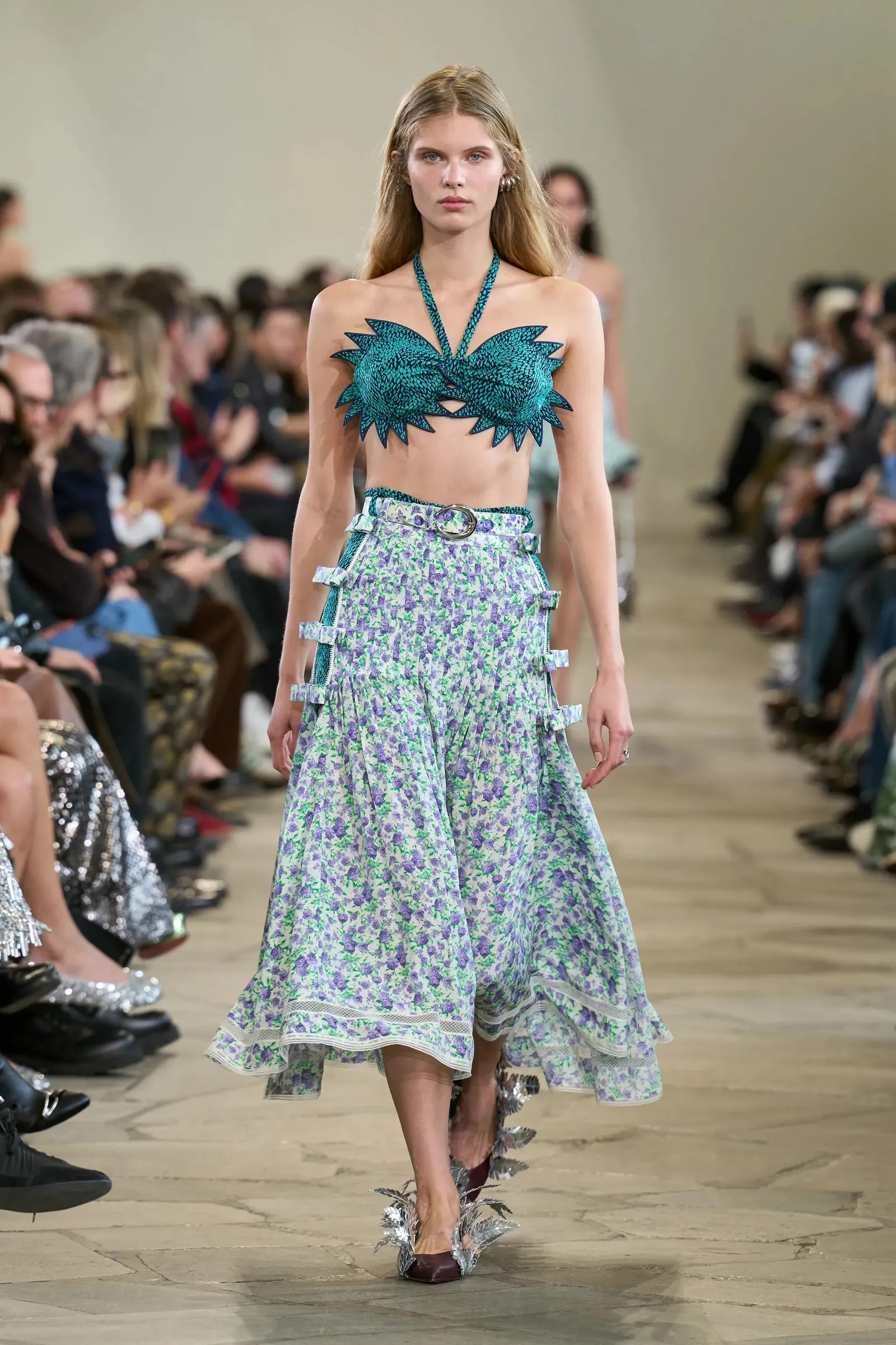 Modelo con top de tirantes verde con formas puntiagudas y falda midi floreada en tonos lilas y blancos sobre pasarela.