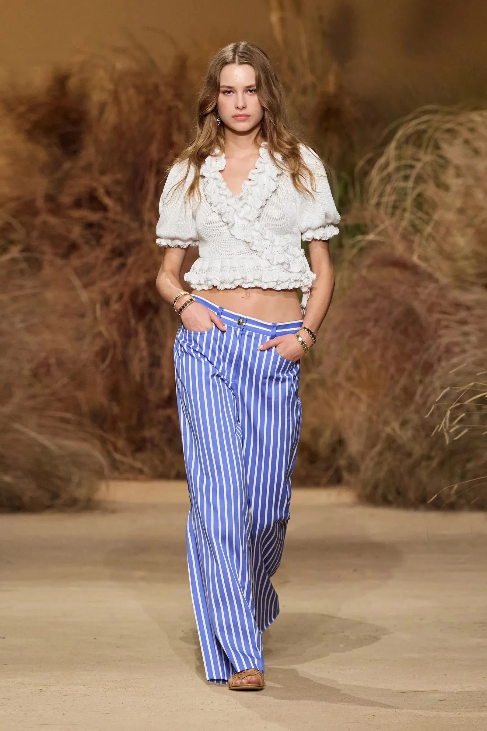 Modelo desfilando con una blusa corta blanca de manga corta con volantes y cuello en V, combinada con pantalones de rayas azules y blancas de talle bajo.