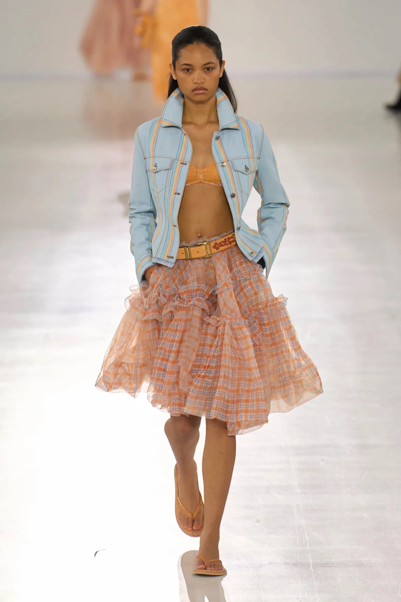 Modelo con chamarra de mezclilla clara, sujetador naranja y falda midi con volantes y cuadros vichy en pasarela.