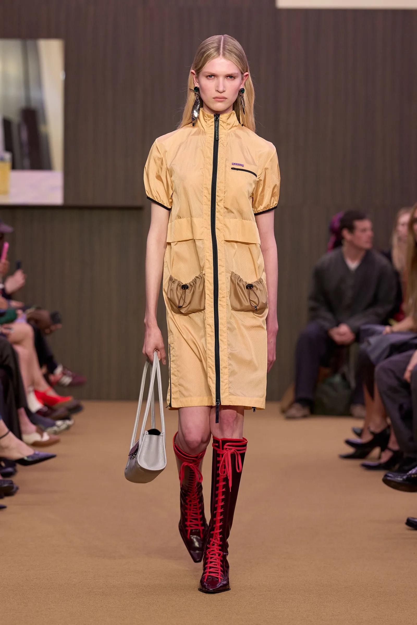 Modelo con vestido utilitario amarillo y botas altas con cordones rojos en pasarela.
