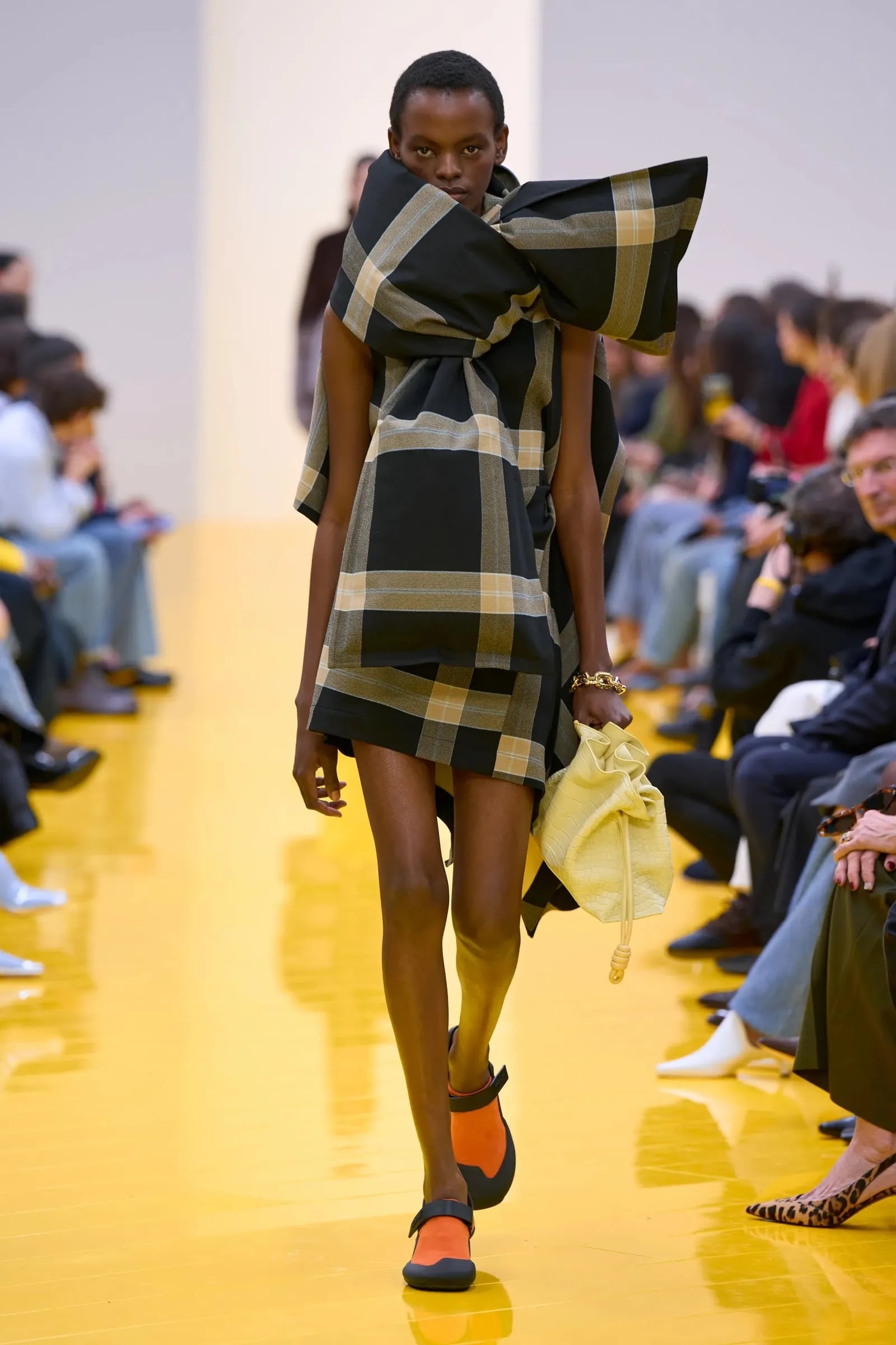 Modelo luciendo un minivestido estructurado de cuadros en tonos negros y beige con un lazo gigante integrado en el cuello, calzado plano y bolso amarillo.