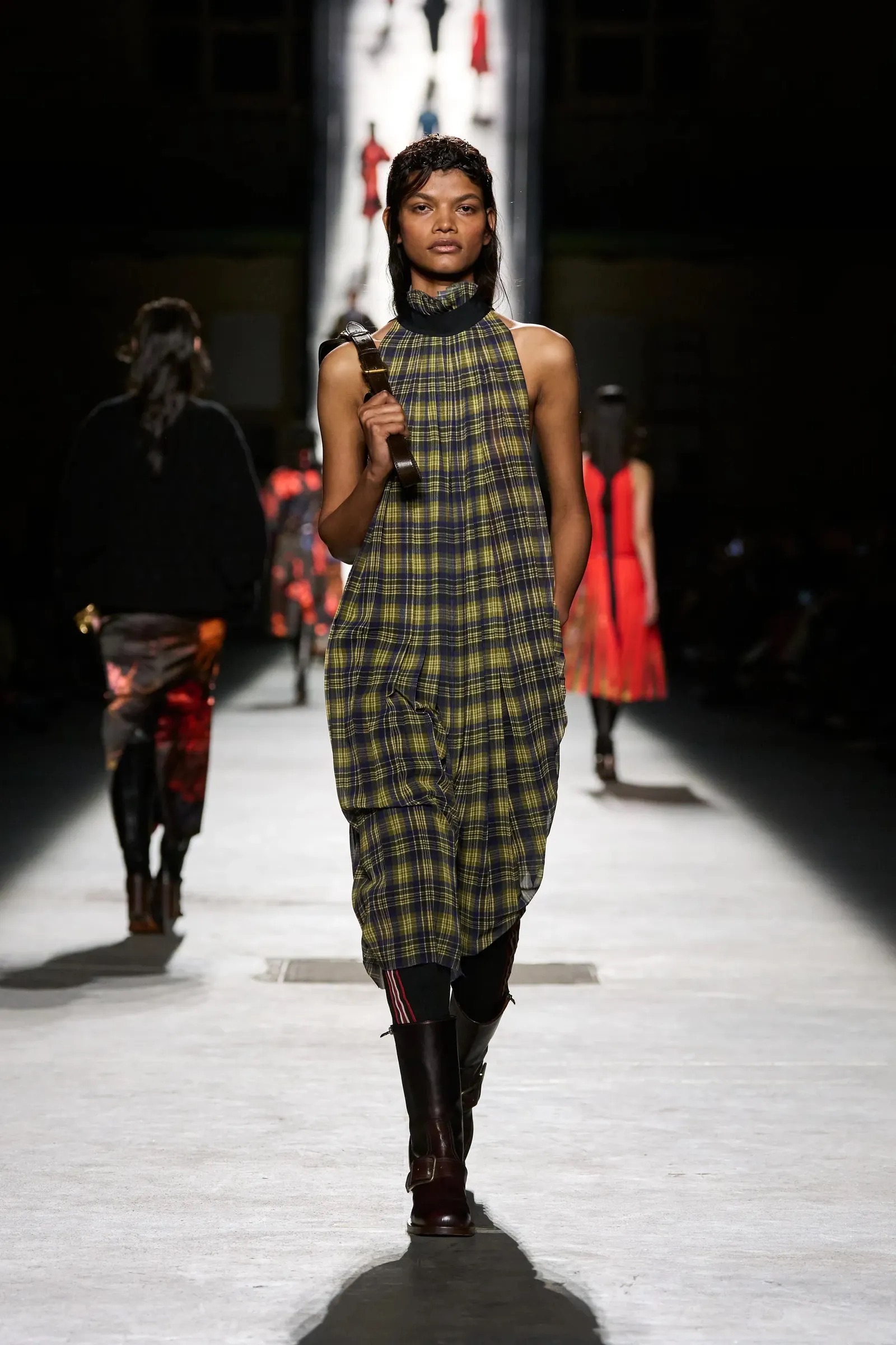 Modelo desfilando con un vestido midi sin mangas de cuadros en tonos verde oliva y amarillo, cuello alto ajustado y botas oscuras de caña alta.