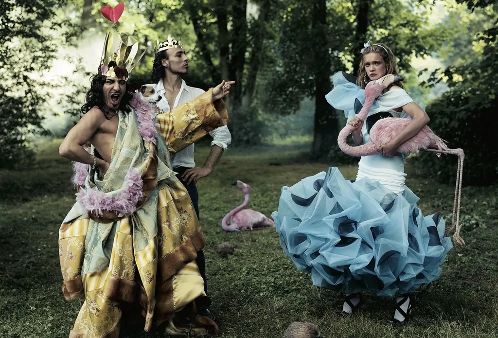 John Galliano disfrazado con una corona y un perro en brazos junto a una modelo sosteniendo flamencos de utilería en un entorno boscoso.