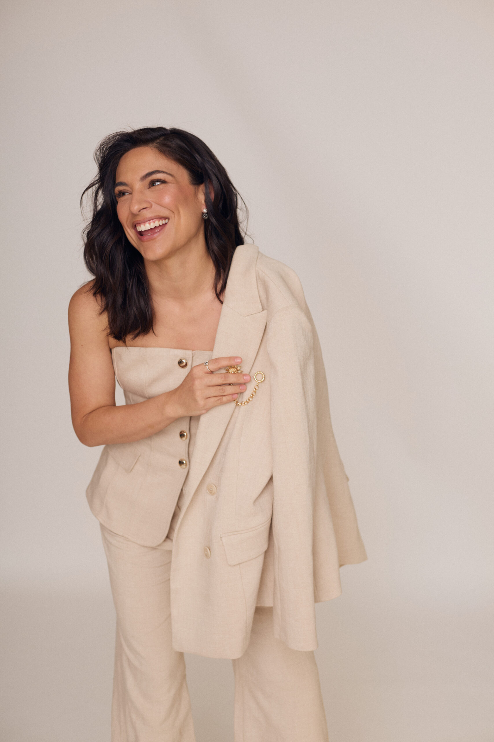 Ana Brenda Contreras sonriendo ampliamente, sujetando la solapa de su saco beige claro decorado con un broche metálico mientras lleva puesto un top ajustado sin tirantes. Styling: JJ Rizo | Maquillaje: Kariana Martínez | Hair: Omar Álvarez