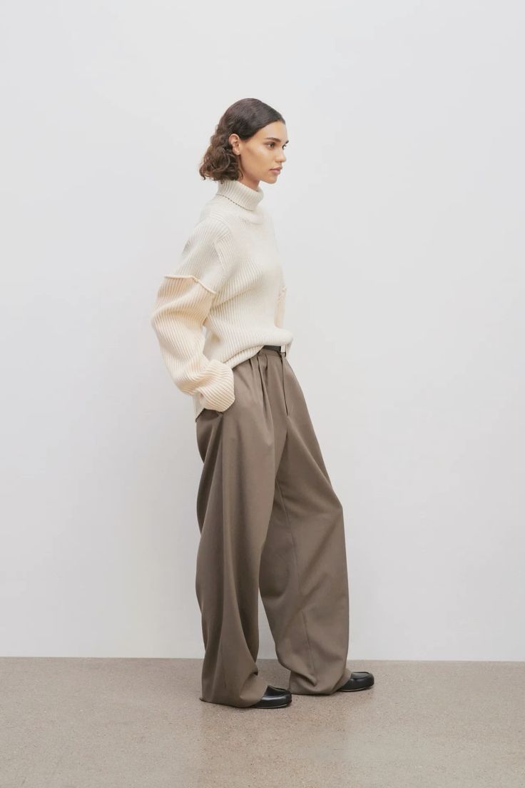 Modelo de perfil luciendo un suéter de punto grueso en tono marfil con cuello alto y mangas amplias, combinado con pantalones de sastre marrones de pernera ancha.