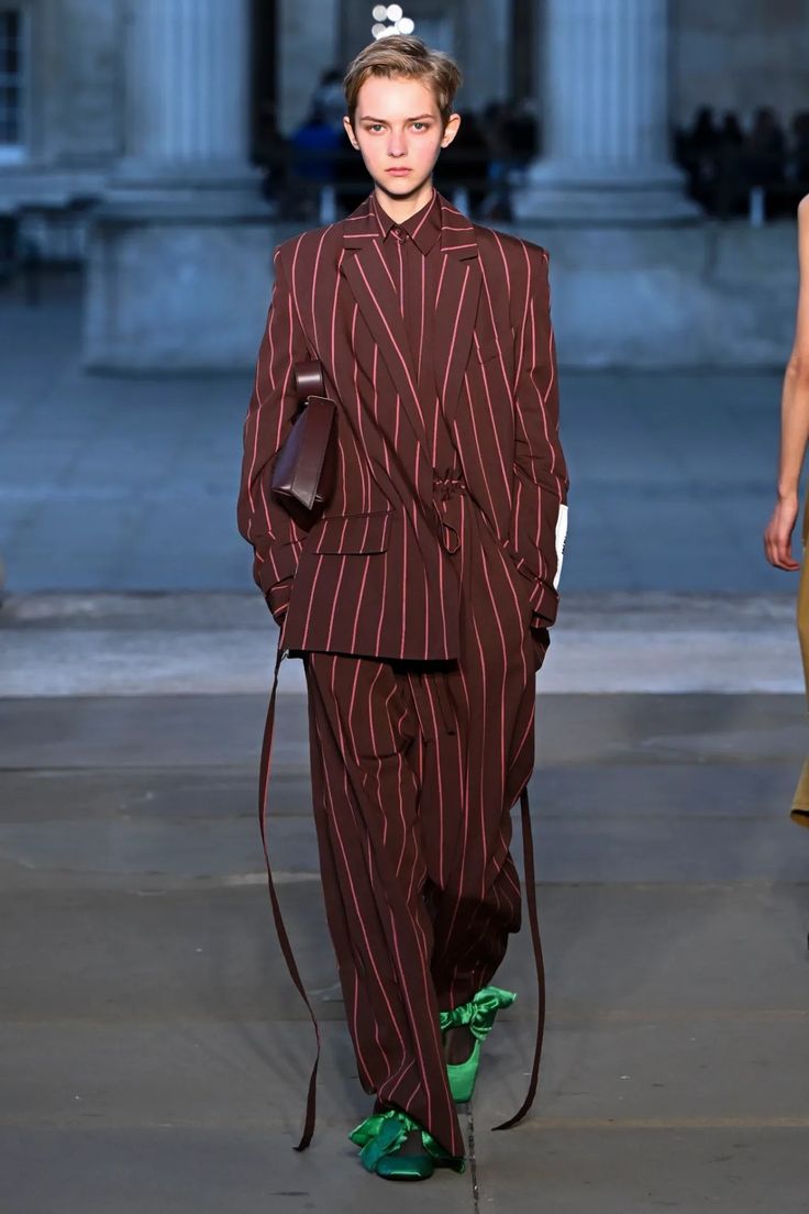 Modelo con un traje sastre estructurado en color marrón con finas rayas verticales (pinstripe) y detalles de lazos verdes en los zapatos.