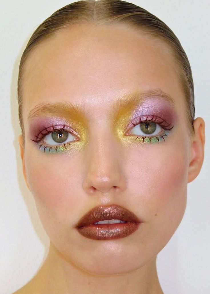 Rostro con maquillaje de ojos detallado utilizando sombras tornasoladas en tonos rosa, amarillo brillante y toques verdosos en las pestañas inferiores, acompañado de labios metalizados.