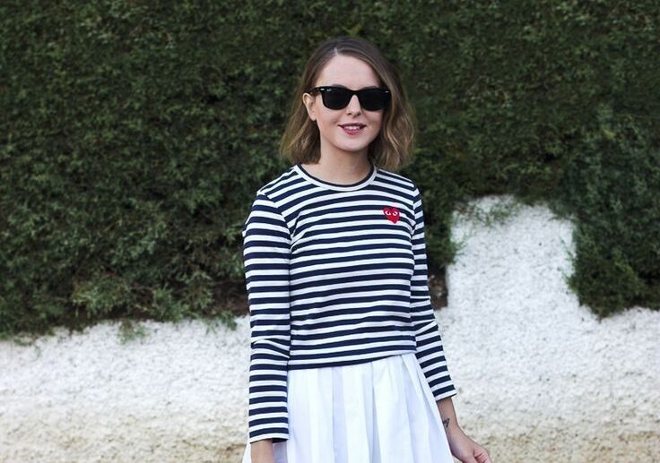 Mujer de pie en la calle vistiendo una blusa de rayas marineras en azul marino y blanco con un pequeño parche de corazón rojo, falda midi blanca de corte amplio, tenis oscuros y bolso negro.
