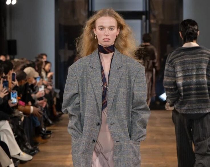 Modelo desfilando con un blazer gris amplio de cuadros, un vestido fluido en tono rosa claro, bufanda fina en el cuello y zapatos oscuros con calcetines marrones.