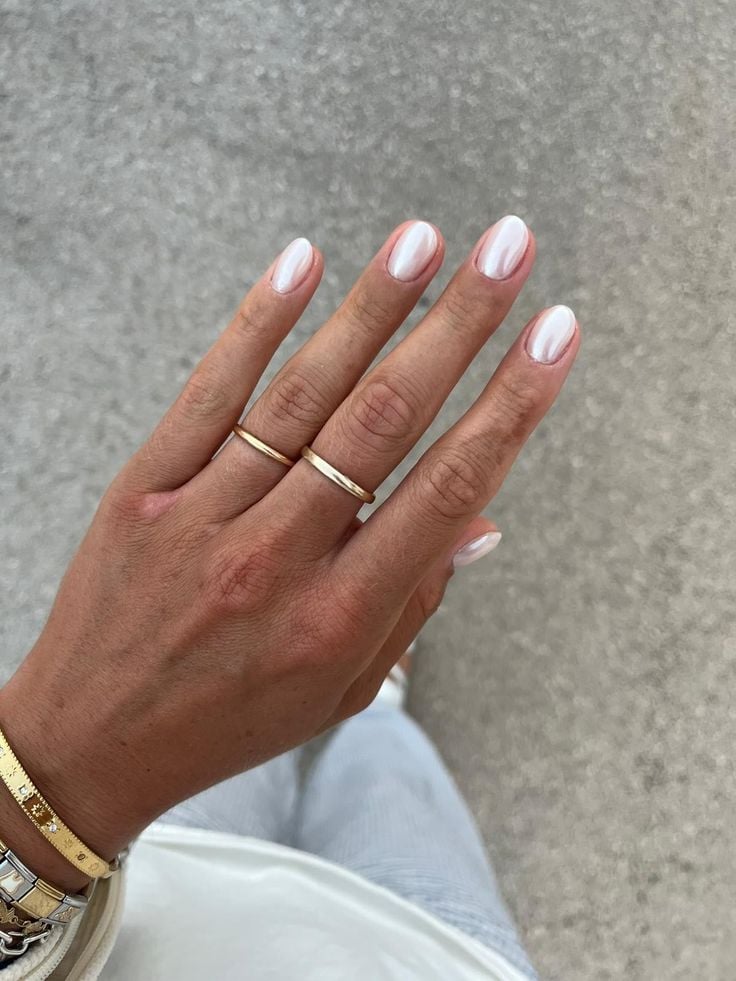Mano con anillos sencillos luciendo uñas cortas almendradas con un acabado de polvo cromado blanco perlado muy luminoso.