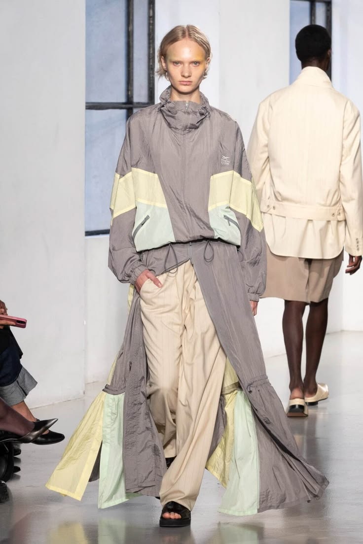 Modelo en pasarela con una chaqueta deportiva impermeable gris con paneles en tonos pastel y una falda larga fluida integrada.