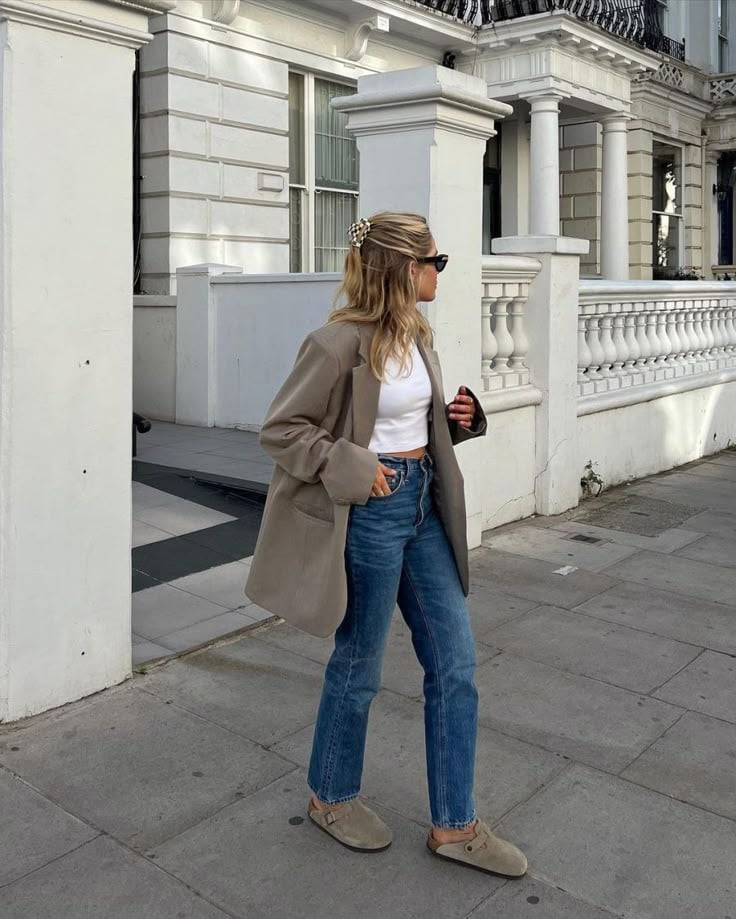 Mujer con blazer taupe, crop top blanco, jeans rectos y zuecos de ante caminando por la calle.