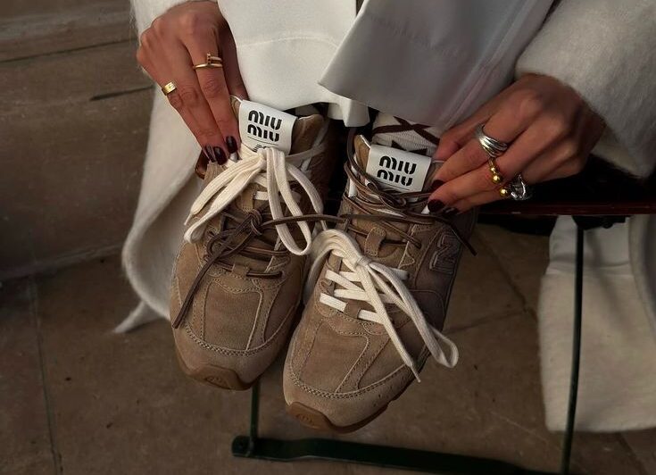 Primer plano de unas manos sosteniendo unos tenis de ante en tono marrón con detalles claros y agujetas blancas, con una chaqueta blanca de textura suave al fondo.