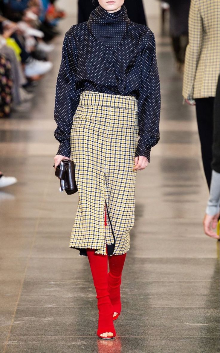 Modelo en pasarela luciendo una blusa de cuello alto en tono oscuro con estampado de lunares blancos, una falda lápiz midi a cuadros amarillos y grises con abertura frontal, y medias rojas con sandalias abiertas.