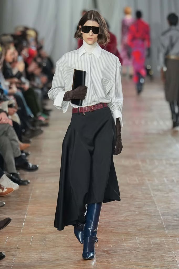 Modelo en pasarela luciendo una camisa de rayas con cuello blanco almidonado, falda midi negra estructurada, cinturón burdeos, gafas de sol oscuras y botas altas azules.