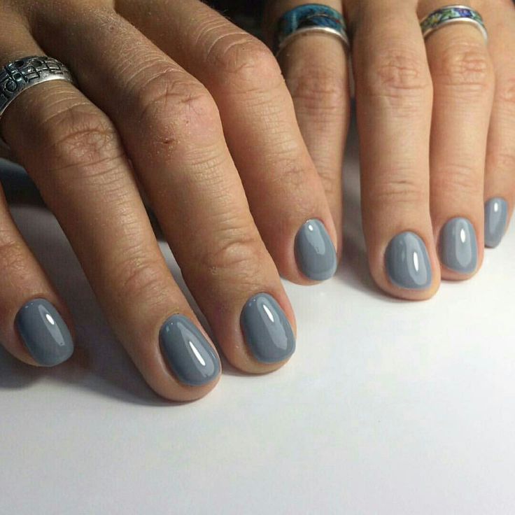 Manos con anillos decorativos luciendo uñas cortas almendradas con un esmalte en tono gris pizarra de acabado brillante.
