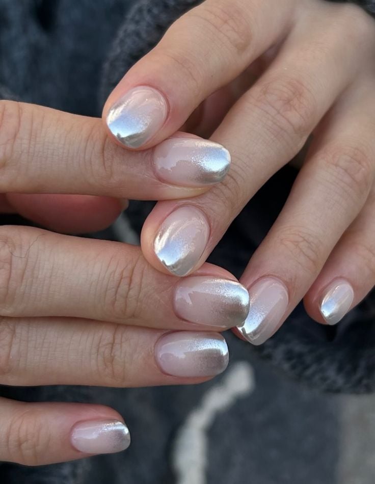 Manos juntas mostrando uñas cortas con un diseño que combina una base neutra y un efecto metálico cromado difuminado en las puntas.