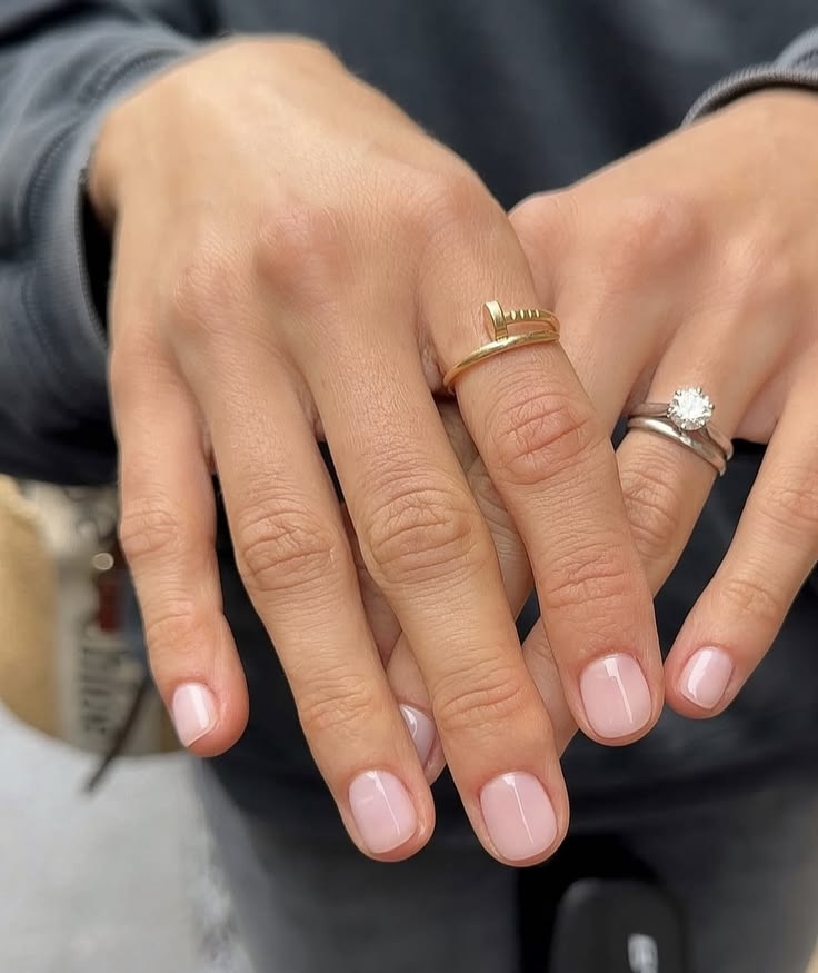 Manos con manicura corta y limpia en un tono rosa muy suave y natural, luciendo un anillo minimalista dorado de clavo y un anillo de compromiso con diamante.