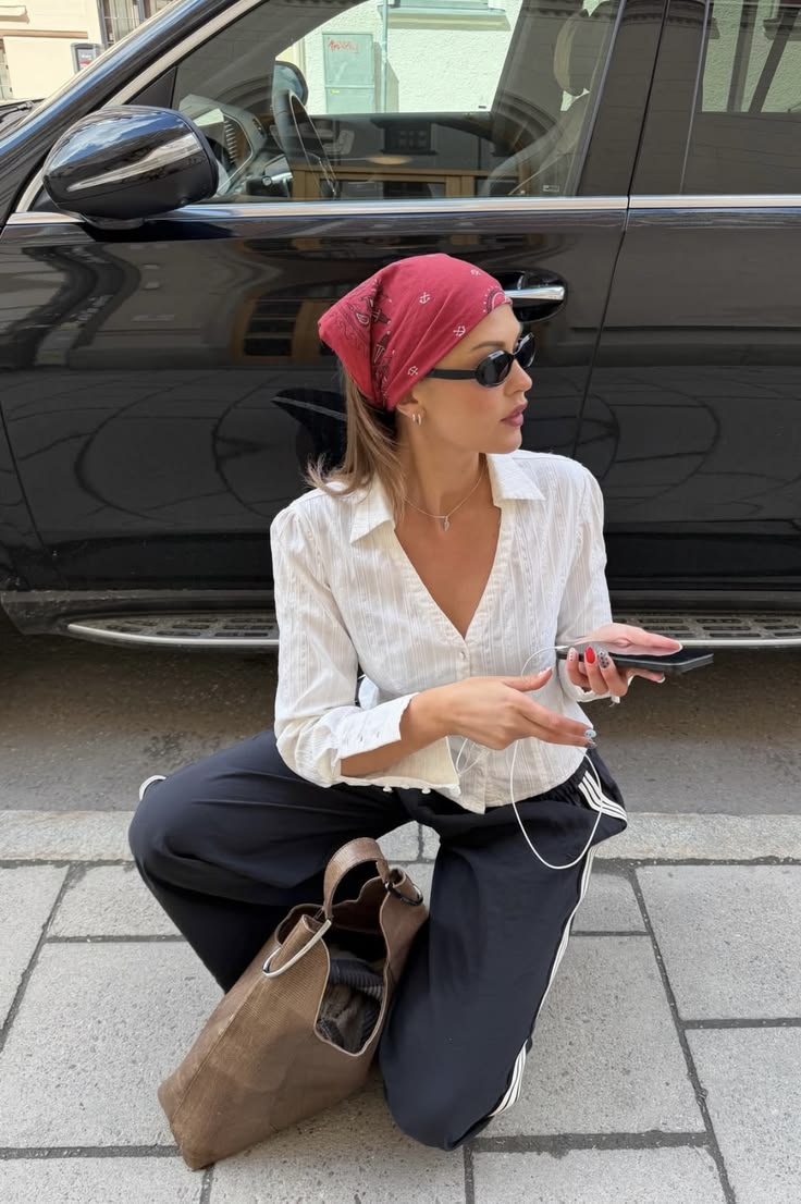 Mujer agachada en la calle con bandana roja, camisa blanca de botones, pantalones deportivos oscuros y bolso marrón