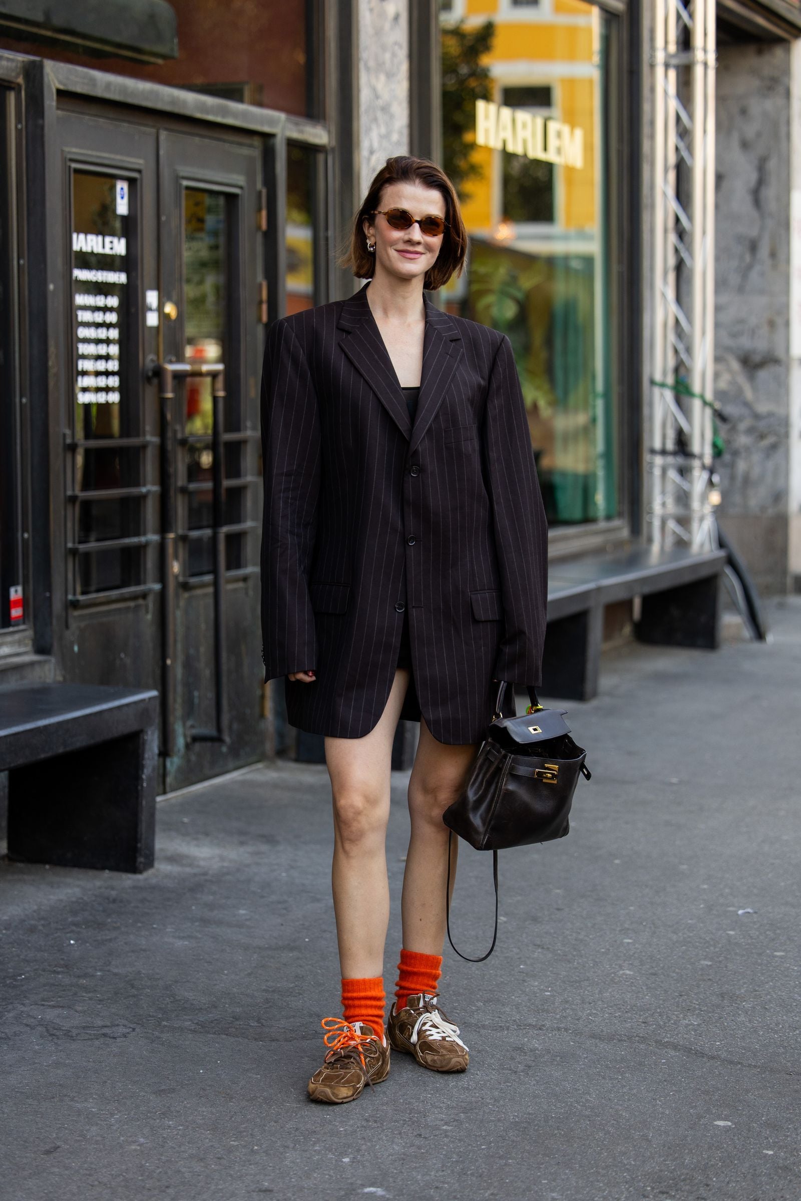 Mujer de pie en la calle luciendo un blazer sastre oversized de rayas diplomáticas, calcetines naranjas vibrantes y tenis de ante marrón.