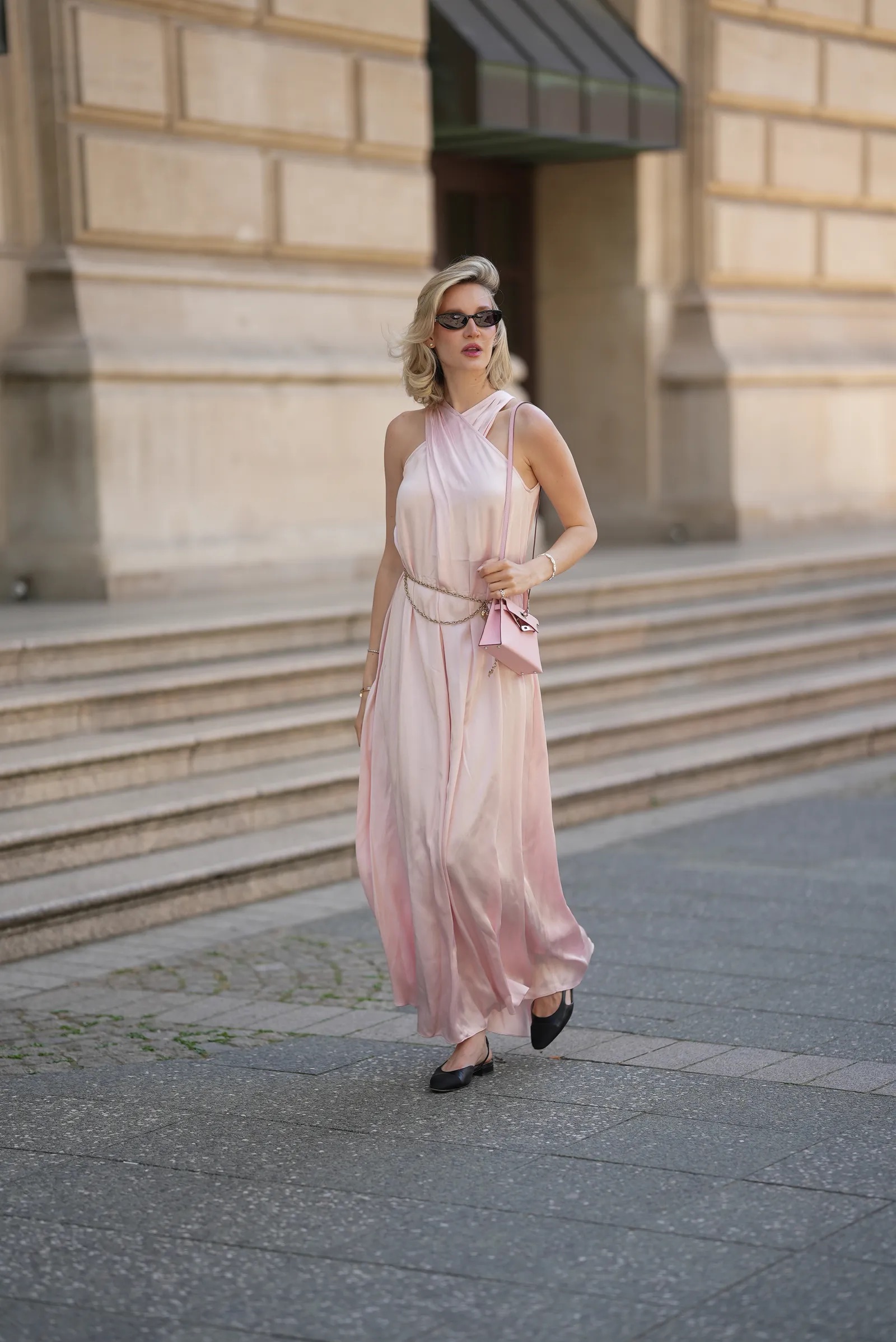Mujer caminando por la calle luciendo un vestido largo satinado de color rosa pálido, gafas de sol y bailarinas slingback negras de diseño plano.