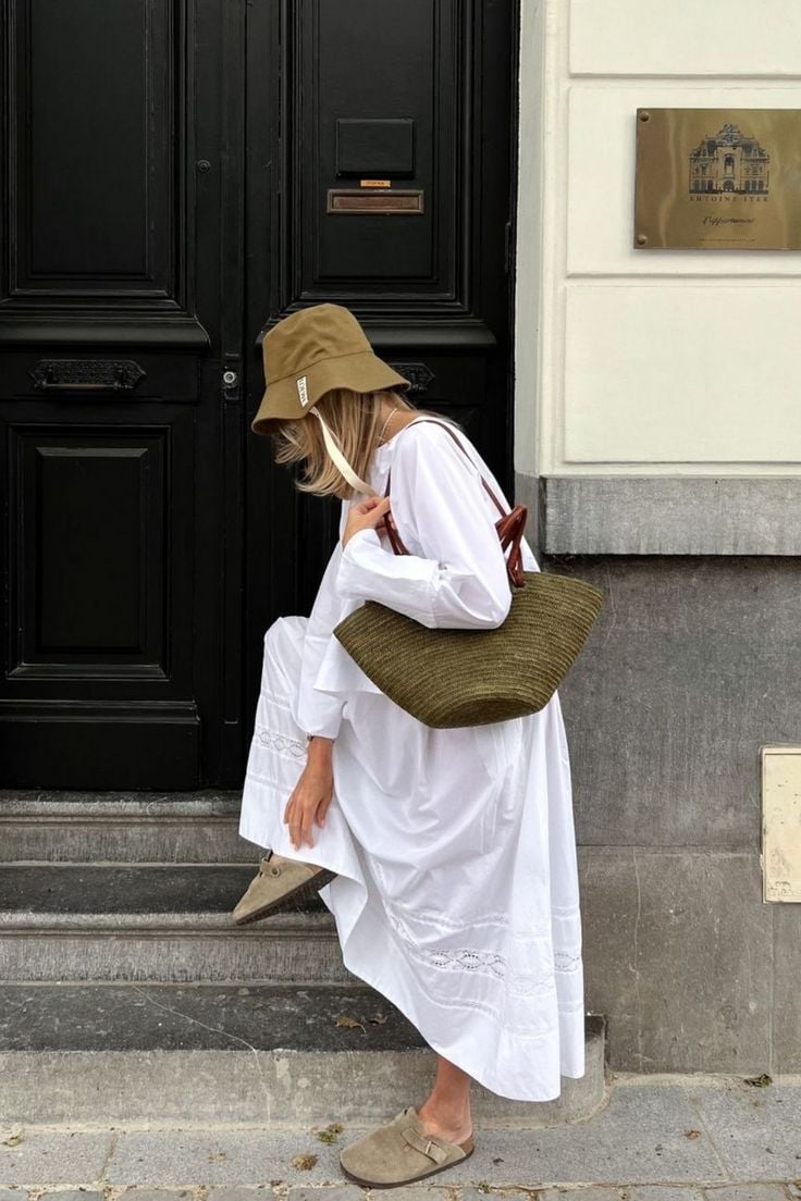 Mujer con vestido blanco largo con detalles bordados, sombrero tipo bucket y zuecos de ante.