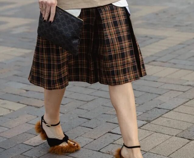 Persona caminando en la calle vistiendo un suéter gris claro metido en una falda midi a cuadros británicos en tonos marrones y negros, con sandalias de tacón decoradas con pelo sintético.