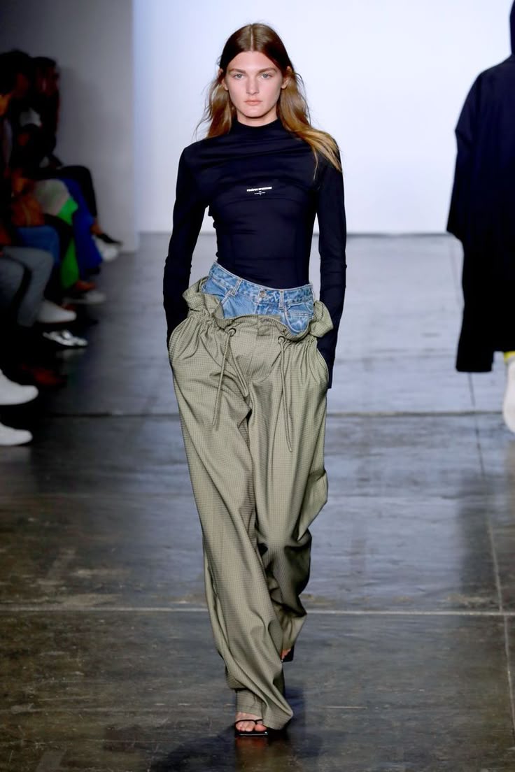 Modelo desfilando con un suéter negro de cuello alto, pantalones anchos en tono verde olivo con jareta y un detalle superpuesto de mezclilla en la cintura.