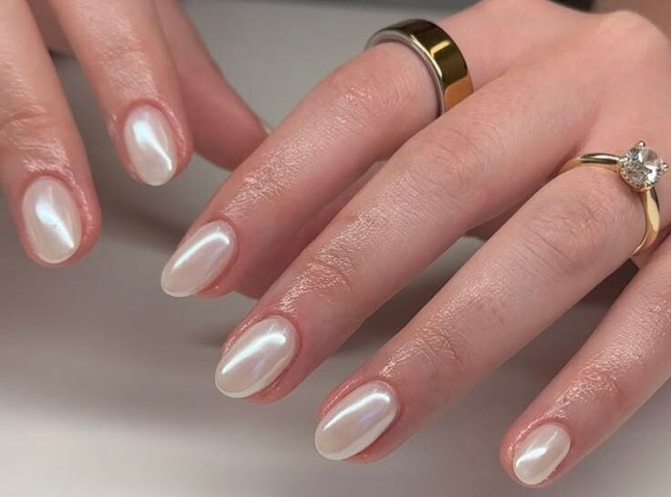 Manos con anillos dorados mostrando uñas ovaladas medianas con un esmalte blanco perlado de intenso brillo nacarado.