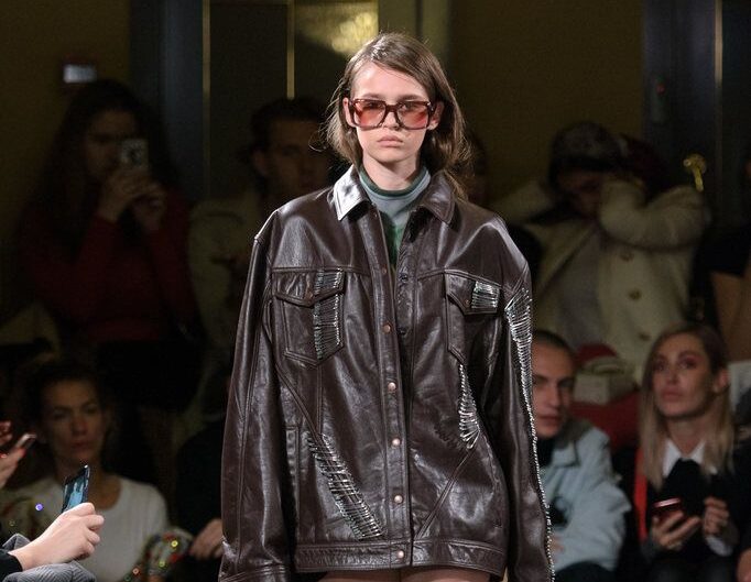 Modelo en pasarela luciendo una chaqueta corta de cuero marrón oscuro decorada con múltiples imperdibles metálicos, combinada con botas altas de piel.