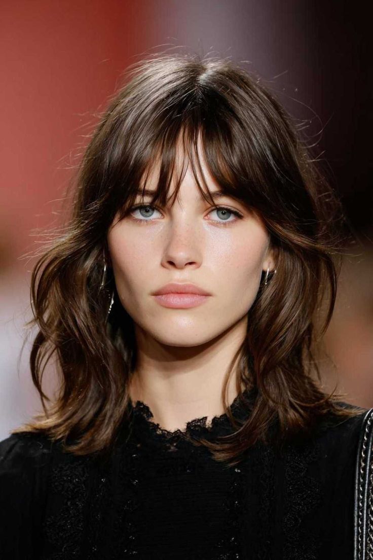  Rostro de una modelo con cabello castaño oscuro con ondas suaves, corte lob y flequillo desfilado abierto sobre la frente.