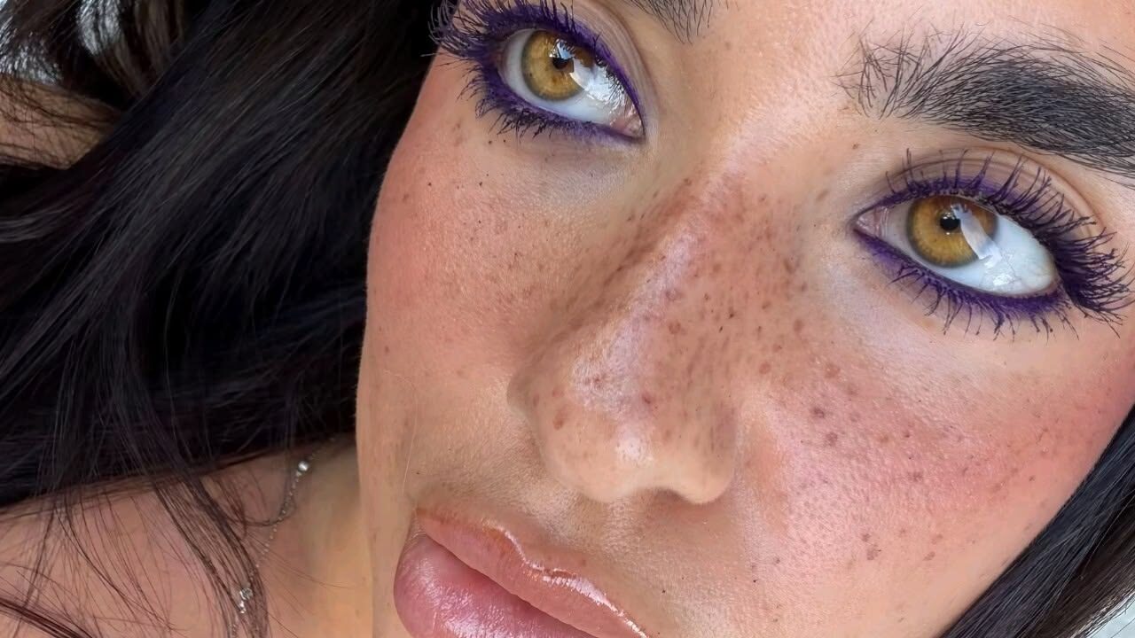 Primer plano de un rostro con pecas marcadas, ojos color avellana con máscara de pestañas en un tono morado profundo enmarcando la mirada.