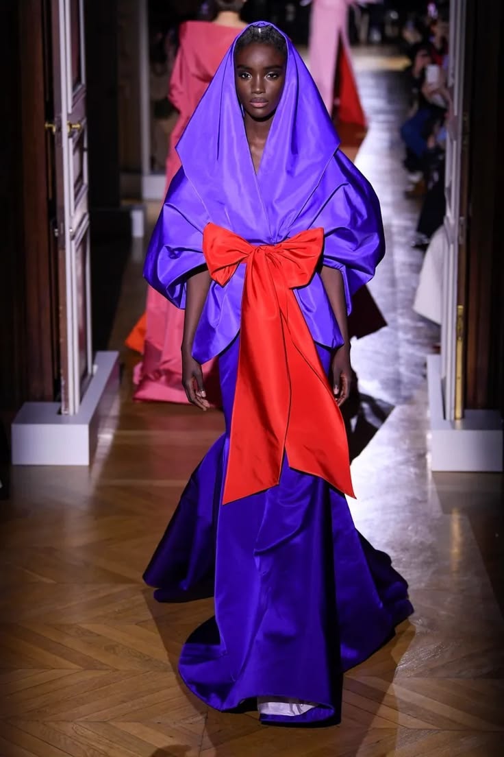 Modelo en desfile portando un vestido estructurado de alta costura en color morado brillante con una capucha integrada y un gran lazo rojo en la cintura.
