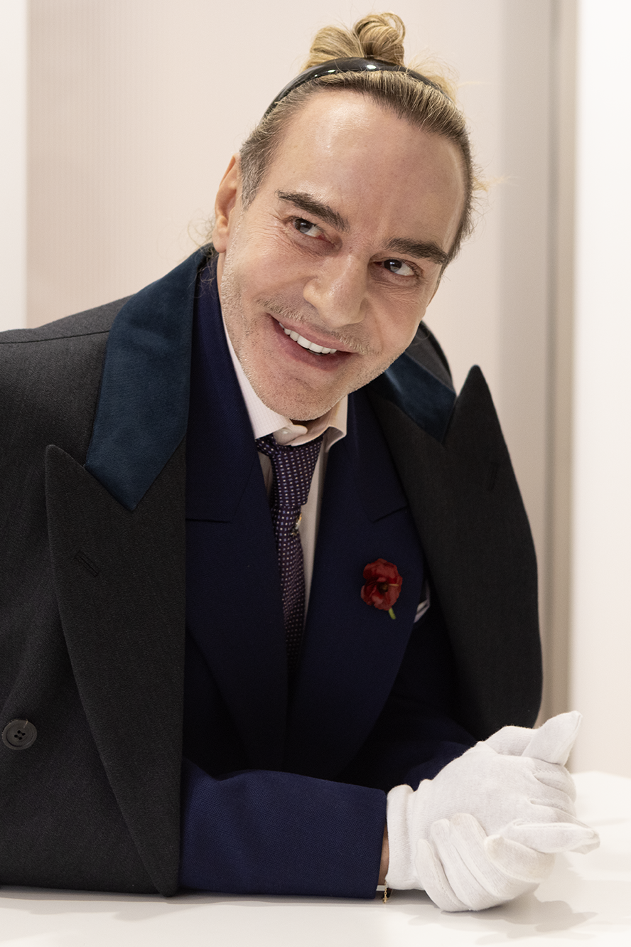Retrato en primer plano de John Galliano sonriendo, vistiendo abrigo oscuro con solapas azules, corbata estampada y guantes blancos de atelier.