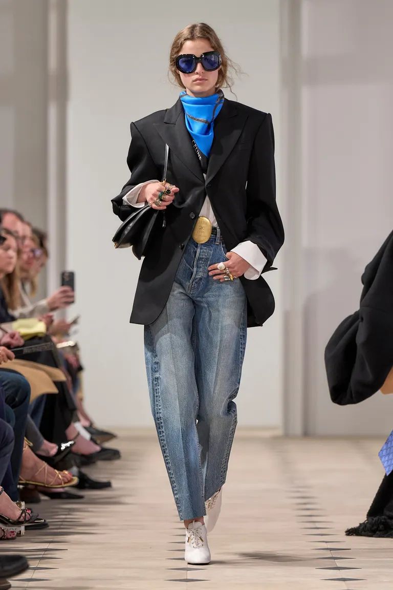 Modelo en pasarela con un blazer negro oversize, pañuelo azul al cuello, gafas de sol y jeans clásicos de mezclilla azul con pinzas