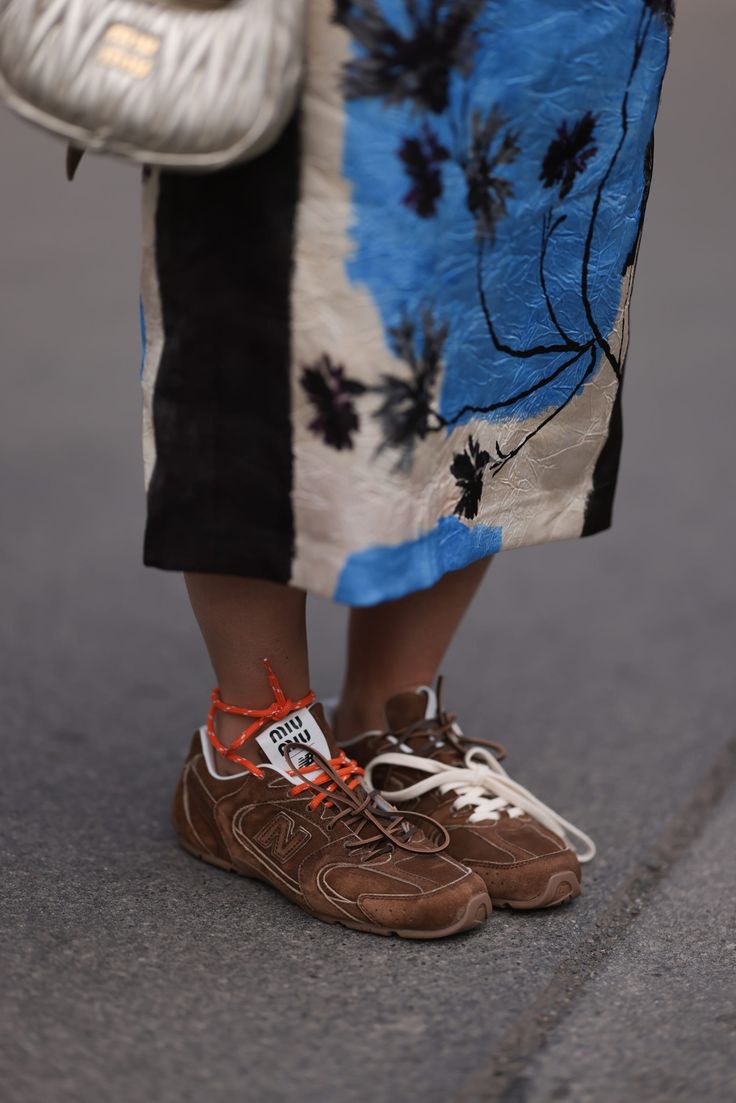 Detalle de las piernas de una persona vistiendo una falda con estampado azul y negro, calcetines naranjas y tenis de ante marrón con cordones claros.