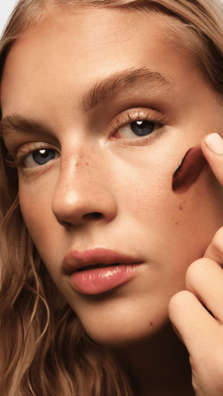 Primer plano de una modelo aplicando un producto bronceador en crema en tono cálido sobre la mejilla con la yema del dedo