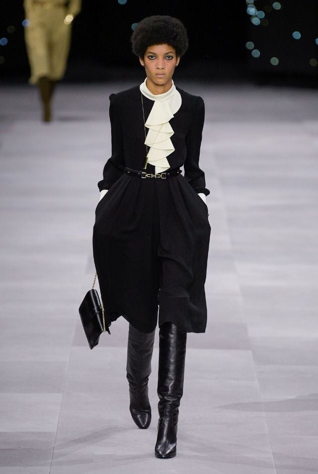 Modelo con cabello corto luciendo un conjunto negro de bermudas amplias de talle alto, una blusa con cuello alto de olanes blancos prominentes y botas altas de piel negra.