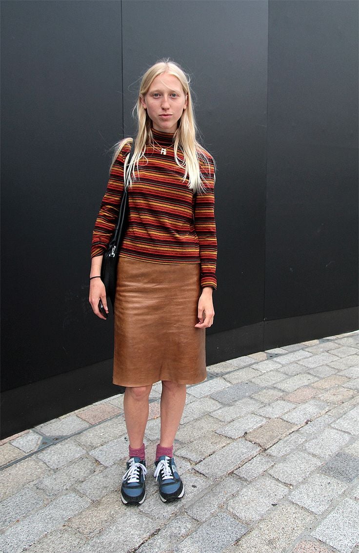 Mujer con cabello rubio lacio luciendo una blusa de cuello alto con rayas multicolores, una falda lápiz de piel marrón, calcetines rosas y tenis deportivos oscuros.