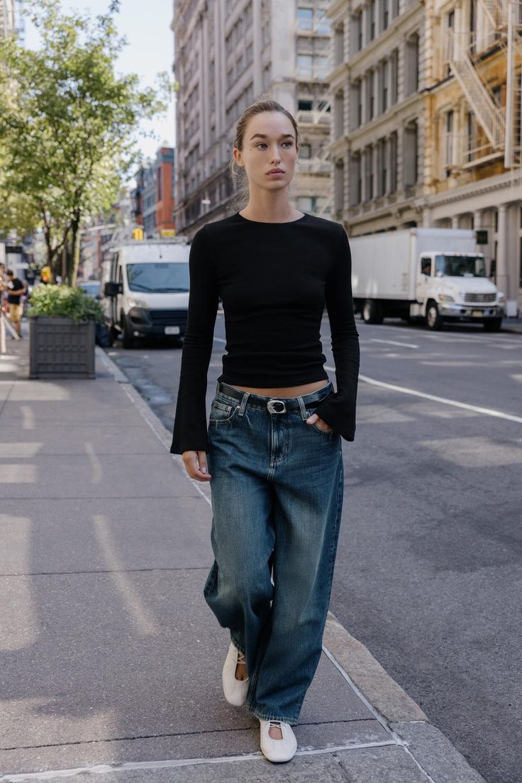 Modelo caminando por la calle con una camiseta negra entallada de manga larga acampanada, vaqueros anchos de tiro medio y bailarinas blancas.