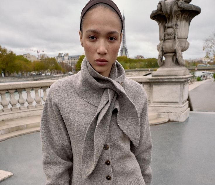 Modelo de pie en un puente luciendo una chaqueta de punto gris con cuello integrado en forma de pañuelo desatado, falda marrón y guantes sin dedos.