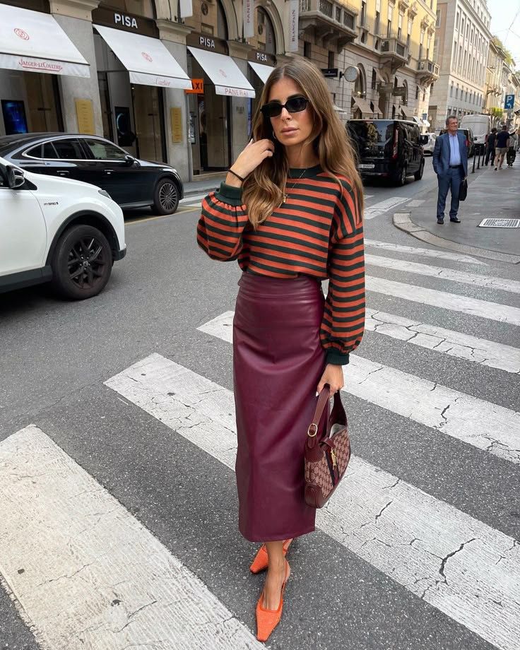 Mujer cruzando la calle con un suéter de rayas en tonos oscuros, falda lápiz larga de piel en color burdeos, bolso de mano estampado y zapatos de tacón naranja.